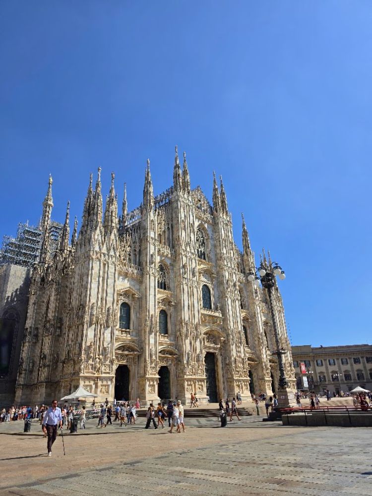 Milano katedros Duomo di Milano gotikinis fasadas su daugybe smailių bokštelių ir puošniomis akmens skulptūromis. Priešais katedrą saulėtą dieną vaikštinėja turistai, o virš jos driekiasi giedras mėlynas dangus.